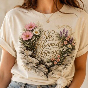 Può includere: Una t-shirt color crema con una grafica di pietra incrinata a forma di cuore, riempita di fiori selvatici rosa, bianchi e viola. Il testo dorato in corsivo al centro recita "She Bloomed Anyway". La t-shirt ha le maniche corte.