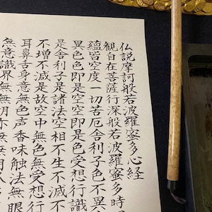 以下が含まれることがあります： オフホワイトの紙に、黒い伝統的な中国の書道文字が縦列で書かれています。紙の横には、明るい色の木製ハンドルが付いた細長い書道用筆と、金色の龍の形をした筆置きが置かれています。