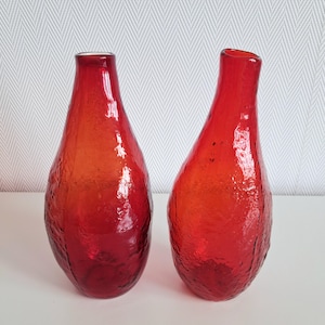 Pode incluir: Dois vasos de vidro vermelho correspondentes com uma forma orgânica e assimétrica distinta. O vidro apresenta um acabamento de superfície texturizado e granulado que capta a luz, enquanto os gargalos são lisos e estreitos, para uma base arredondada.