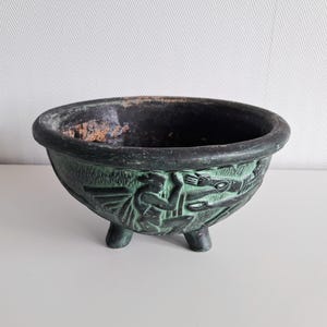 Puede incluir: Un cuenco de color verde oscuro y negro con tres patas cortas y un borde redondeado. El exterior presenta un diseño en relieve que representa figuras estilizadas en movimiento. El interior del cuenco es oscuro y muestra signos de desgaste con una superficie texturizada.