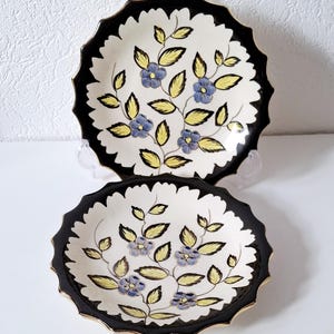 Könnte beinhalten: Zwei runde Keramikteller mit gewelltem Rand, die eine cremefarbene Mitte mit handgemalten blauen Blumen und gelben Blättern aufweisen. Der breite, schwarze, gewellte Rand ist mit einem dünnen Goldrand an der Außenkante versehen.