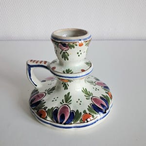 Può includere: Un portacandele in ceramica con una base circolare ampia e una piccola impugnatura. La superficie bianco sporco è decorata con motivi floreali dipinti a mano nei toni del blu, rosa, verde e arancione, con bordi delineati in blu e finitura lucida.