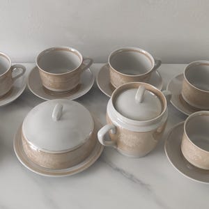 Könnte beinhalten: Ein Teeservice bestehend aus sechs Tassen mit passenden Untertassen, einer Zuckerdose mit Deckel und einem Milchkännchen mit Deckel. Jedes Teil hat ein weißes Inneres und ein hellbeiges Äußeres mit einem dezenten, sich wiederholenden Muster.