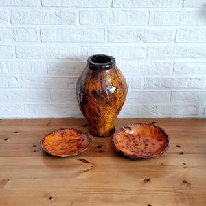 Puede incluir: Un conjunto de cerámica que consta de un jarrón alto y bulboso y dos cuencos circulares poco profundos. Cada pieza presenta un esmalte brillante y moteado en tonos naranja vibrante, ámbar intenso y marrón oscuro, con un acabado liso y reflectante.