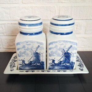 Può includere: Due barattoli in ceramica bianca con coperchi blu e vassoio rettangolare abbinato, ciascuno decorato con una stampa blu e bianca di un paesaggio con mulini a vento. I barattoli hanno una base quadrata e una parte superiore arrotondata.