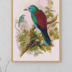 May include: A cross-stitch pattern featuring two birds perched on a tree branch. The larger bird has a bright blue head, a dark red back, and green wings. A smaller, similar bird sits on a thinner branch nearby. The design is set against a light pink background and framed in a light wood frame.