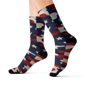 May include: A pair of crew-length socks featuring a camouflage pattern in red, white, and blue. The design incorporates white five-pointed stars scattered throughout the fabric, with solid black fabric at the toe and heel sections of the socks.