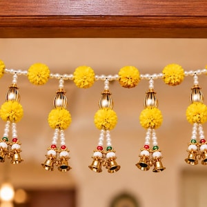 Puede incluir: Un colgante decorativo para puerta que presenta una guirnalda horizontal de flores de caléndula amarillas y perlas blancas artificiales. De la cuerda principal cuelgan borlas verticales con flores amarillas, perlas y campanillas doradas.