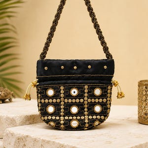 Peut inclure: Un sac à main noir à cordon de serrage doté d'une anse tressée, de broderies dorées et d'accents en miroir circulaires disposés en grille. Le sac comprend des cordons clairs avec des pompons décoratifs dorés de chaque côté.