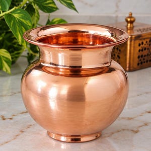 May include: A polished copper pot with a rounded body, a wide neck, and a flared rim. The vessel features a smooth, reflective metallic surface with a warm reddish-brown tone. It has a circular base and a consistent, uniform finish throughout.