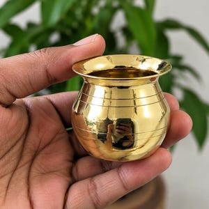 May include: A small, polished gold-colored metal pot with a wide, flared rim and a rounded body. The surface of the vessel features several thin, horizontal engraved lines circling the exterior, creating a simple, reflective, and smooth finish.
