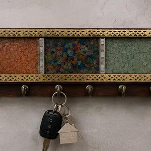 May include: A rectangular wall-mounted key holder featuring a dark wood base with six metal hooks. The upper section has a gold-toned metal frame with a lattice pattern, enclosing three panels filled with orange, multicolored, and green crushed stones.