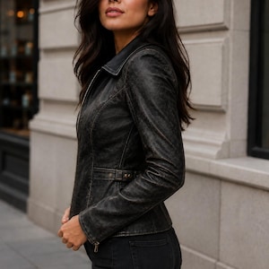 May include: A black leather jacket with a distressed finish, featuring a classic collar, a full-length front zipper, long sleeves with zippered cuffs, and a fitted waist with side adjustment tabs, paired with black denim jeans.
