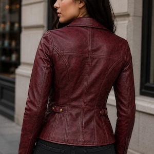 May include: A deep burgundy leather jacket featuring a structured collar, long sleeves with buttoned cuffs, and a fitted waist with side adjustment tabs. The jacket has visible vertical seam detailing across the back and is worn over black pants.