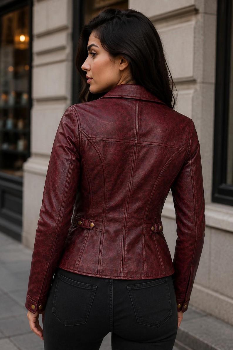 May include: A deep burgundy leather jacket featuring a structured collar, long sleeves with buttoned cuffs, and a fitted waist with side adjustment tabs. The jacket has visible vertical seam detailing across the back and is worn over black pants.
