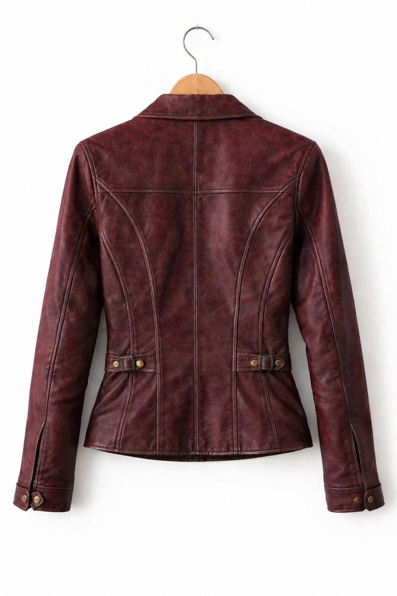 May include: A dark burgundy jacket with a distressed finish, featuring a collared neckline, long sleeves with buttoned cuffs, and adjustable side tabs with brass-toned hardware. The back panel includes vertical seam detailing and a structured fit.