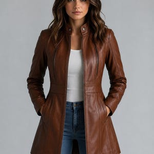 Könnte beinhalten: Eine mittellange braune Lederjacke mit Stehkragen und Druckknopfverschluss, durchgehendem Reißverschluss, vertikalen Ziernähten und zwei Seitentaschen. Die Jacke wird über einem weißen Oberteil und einer blauen Jeans getragen.