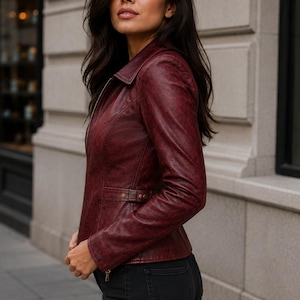 May include: A deep burgundy leather jacket featuring a classic collar, a full-length front zipper, and a fitted silhouette. The jacket includes long sleeves with zippered cuffs and a side waist adjustment strap with metallic hardware details.