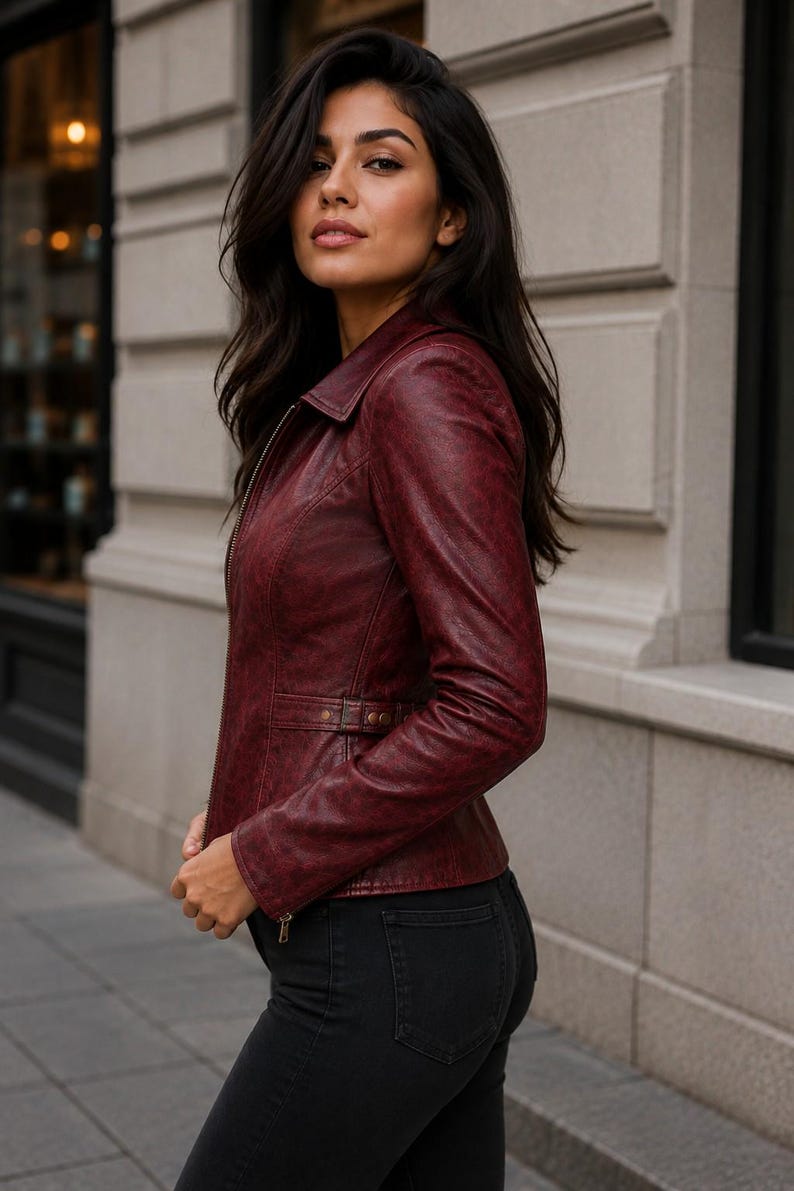May include: A deep burgundy leather jacket featuring a classic collar, a full-length front zipper, and a fitted silhouette. The jacket includes long sleeves with zippered cuffs and a side waist adjustment strap with metallic hardware details.