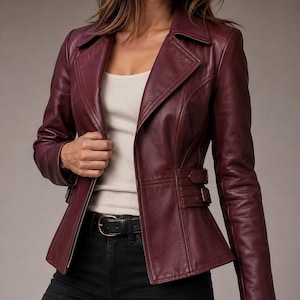 May include: A deep burgundy leather jacket featuring a notched collar, long sleeves, and a fitted waist with two adjustable side buckle straps. The jacket has a front zipper closure and vertical seam detailing that creates a structured silhouette.