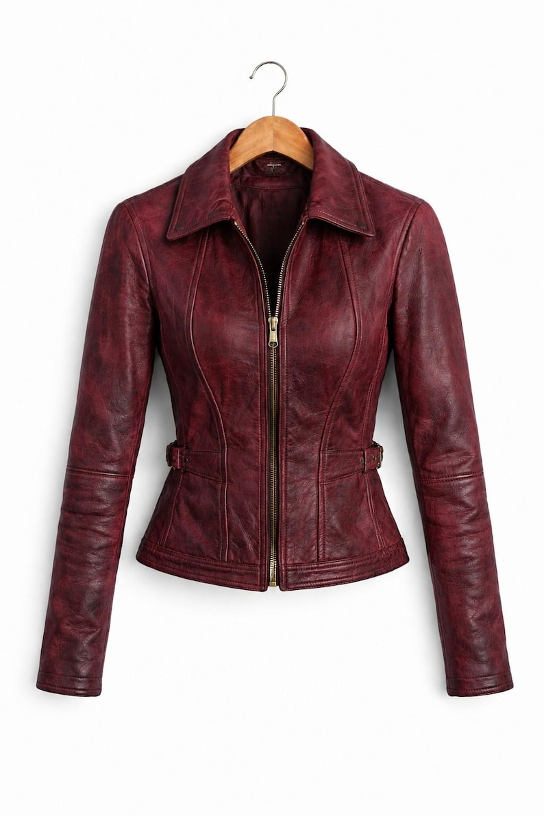 May include: A deep burgundy jacket with a distressed finish, featuring a pointed collar, long sleeves, and a central gold-toned zipper closure. The garment includes vertical seam detailing and adjustable side waist tabs with small buckles.