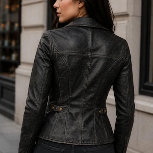 May include: A distressed black leather jacket featuring a structured fit, a pointed collar, and vertical seam detailing. The jacket includes side waist tabs with metallic buttons and matching button cuffs, providing a tailored silhouette.