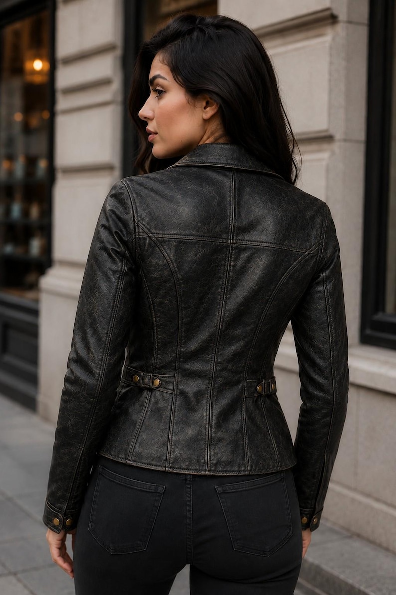 May include: A distressed black leather jacket featuring a structured fit, a pointed collar, and vertical seam detailing. The jacket includes side waist tabs with metallic buttons and matching button cuffs, providing a tailored silhouette.