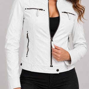 May include: A white jacket featuring a stand-up collar, a central front zipper, and multiple zippered pockets on the chest and sides. The garment includes dark hardware accents, including snaps on the collar and hem, and long sleeves with cuffs.