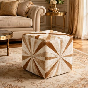 May include: A square ottoman featuring a hair-on-hide texture with a radiating sunburst pattern in alternating shades of cream, beige, and brown. The cube-shaped piece displays a geometric design that converges at the centre of each visible side.
