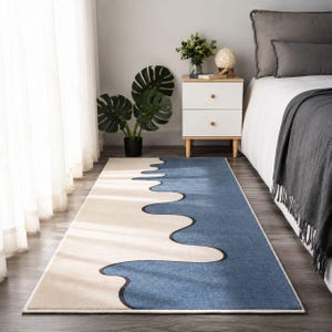 K&ouml;nnte beinhalten: Ein rechteckiger Teppich mit einem zweifarbigen Design, bestehend aus einem cremefarbenen und einem blauen Bereich, die durch eine gro&szlig;e, flie&szlig;ende, wellenf&ouml;rmige Linie getrennt sind, welche diagonal &uuml;ber die Oberfl&auml;che verl&auml;uft.