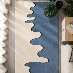 K&ouml;nnte beinhalten: Ein rechteckiger Teppich mit einem zweifarbigen Design, bestehend aus einem cremefarbenen und einem blauen Bereich, die durch eine markante, wellenf&ouml;rmige schwarze Linie getrennt sind, welche vertikal durch die Mitte verl&auml;uft.