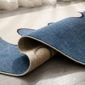 K&ouml;nnte beinhalten: Ein rechteckiger Teppich mit einem Muster aus geschwungenen schwarzen Linien auf blauem und cremefarbenem Grund. Der Teppich ist umgeschlagen und zeigt eine strukturierte blaue Floroberfl&auml;che sowie eine gewebte beigefarbene R&uuml;ckseite mit Kante.