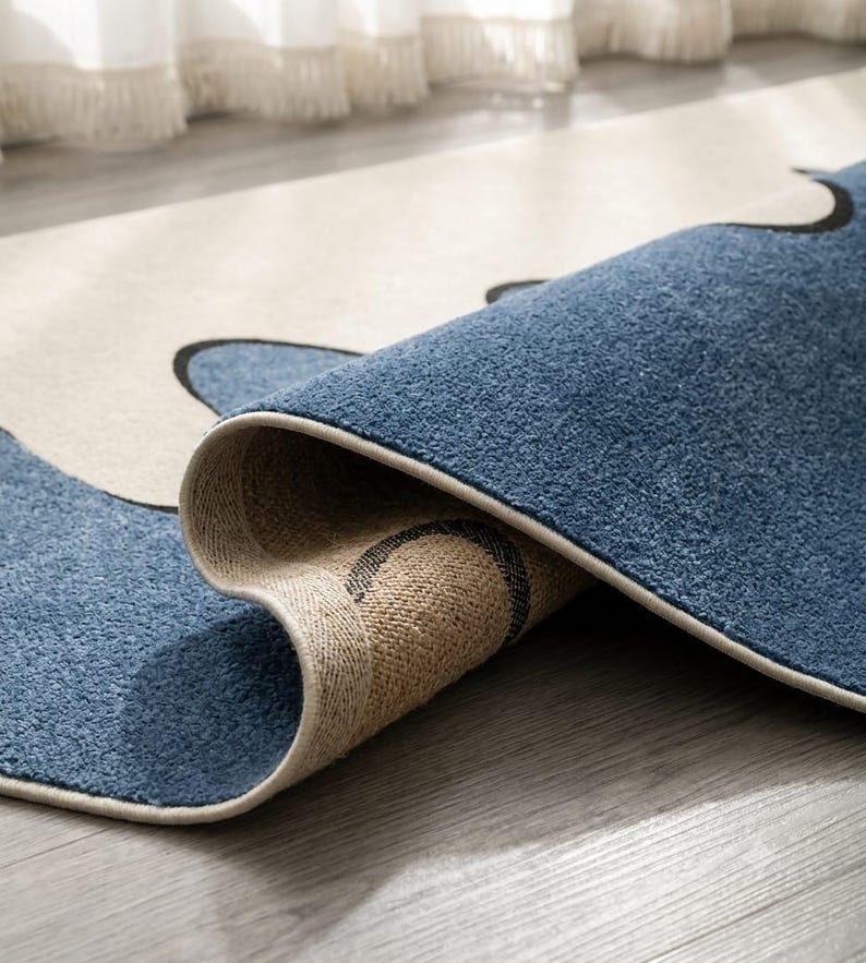 K&ouml;nnte beinhalten: Ein rechteckiger Teppich mit einem Muster aus geschwungenen schwarzen Linien auf blauem und cremefarbenem Grund. Der Teppich ist umgeschlagen und zeigt eine strukturierte blaue Floroberfl&auml;che sowie eine gewebte beigefarbene R&uuml;ckseite mit Kante.
