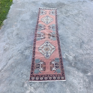 Könnte beinhalten: Ein langer, schmaler Läufer mit einem altrosa Hintergrund und sich wiederholenden geometrischen Rautenmustern in Creme, Dunkelblau und Türkis. Der Teppich ist von einer dunkelblauen Bordüre mit weißem Muster und weißen Fransen umrandet.