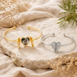 May include: Two open cuff bracelets. The gold-tone bracelet features a central heart-shaped locket with angel wings, displaying a photograph of a family. The silver-tone bracelet features a central heart-shaped charm with angel wings and clear stones.