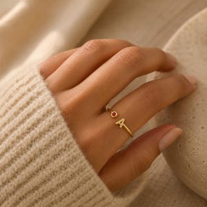 Op de afbeelding: Een goudkleurige open ring met een kleine, ronde, rode edelsteen aan het ene uiteinde en de hoofdletter A aan het andere uiteinde. De ringband is dun en van metaal, ontworpen om aan een vinger te dragen, met een eenvoudige en strakke uitstraling.