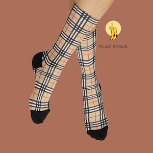 Könnte beinhalten: Wadenhohe Socken mit beiger Basis und einem schwarz-weiß-roten Karomuster. Die Socken haben einfarbig schwarze Fersen und Zehenbereiche. Ein kreisförmiges Logo mit Kleiderbügel-Symbol und dem Text PLAID SOCKS ist oben rechts zu sehen.