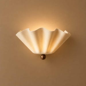 May include: A wall sconce featuring a cream-colored, scalloped, fan-shaped shade that emits a warm, soft glow. The fixture is anchored at the bottom by a small, spherical, dark bronze-toned metal accent piece centered beneath the shade.