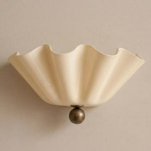 Peut inclure: Une applique murale composée d'un large abat-jour en verre crème à bords festonnés et ondulés. L'abat-jour est fixé sur une base circulaire en métal bronze foncé qui s'étend depuis le centre de la courbe inférieure.