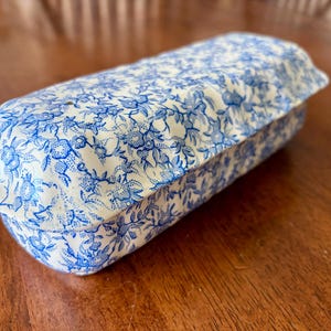 May include: A rectangular ceramic butter dish with a lid, featuring a white base color and an intricate blue floral pattern covering the entire exterior surface. The dish has a slightly scalloped edge along the lid and a smooth, glossy finish.
