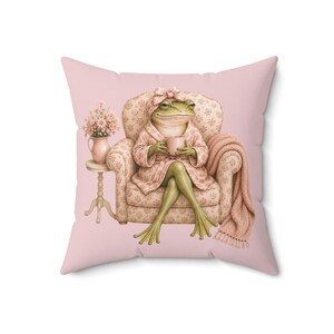 May include: A square throw pillow with a light pink background featuring an illustration of a green frog wearing a floral robe and headwrap, sitting in a floral armchair while holding a mug, next to a small table with a vase of pink flowers.