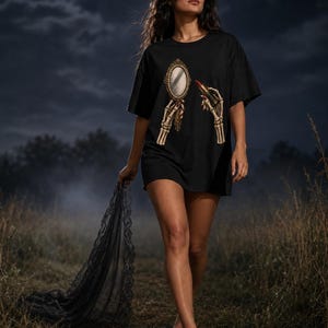 Op de afbeelding: Een zwart t-shirt met een grafische print van twee skeletachtige handen, waarvan er één een versierde handspiegel met een gouden lijst vasthoudt en de andere een tube rode lippenstift. Het ontwerp is gecentreerd op de borst.