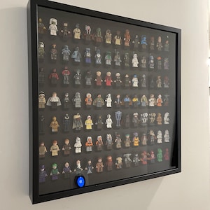 May include: A black rectangular display case with a glass front containing a collection of numerous small plastic character figures arranged in rows. The figures feature various colors and designs, and a small circular blue light is at the bottom.