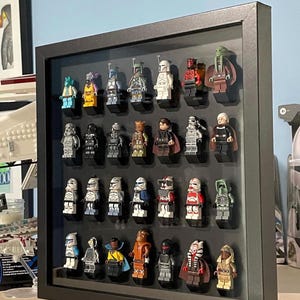May include: A black shadow box frame containing twenty-eight Star Wars themed plastic minifigures arranged in four rows of seven. The figures feature various characters with distinct helmets, capes, and colored outfits mounted on individual black bricks.