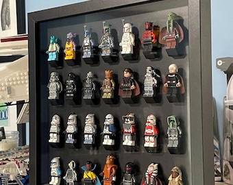 Könnte beinhalten: Ein schwarzer Schattenfugenrahmen mit achtundzwanzig Star Wars-Minifiguren aus Kunststoff, angeordnet in vier Reihen zu je sieben Stück. Die Figuren zeigen verschiedene Charaktere mit markanten Helmen, Umhängen und farbigen Outfits auf schwarzen Steinen.