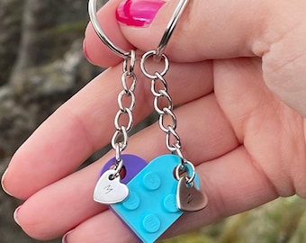 Könnte beinhalten: Zwei ineinandergreifende herzförmige Schlüsselanhänger aus violetten und hellblauen Kunststoff-Bausteinen. Jeder Anhänger besteht aus einer silberfarbenen Metallkette, einem Schlüsselring und einem kleinen Herzanhänger mit den Buchstaben M und Z.