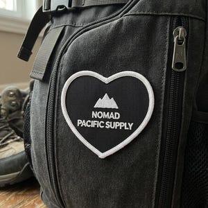 May include: A charcoal gray canvas backpack featuring a heart-shaped patch with a white border. The patch displays a white mountain icon above the text NOMAD PACIFIC SUPPLY. The backpack includes a black plastic buckle and a metal zipper pull.