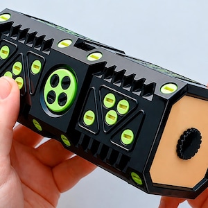 Può includere: Un dispositivo meccanico rettangolare nero con accenti circolari e triangolari verde lime brillante. La struttura include un pannello circolare centrale con quattro fori e un tappo piatto color beige chiaro con una piccola manopola nera testurizzata.