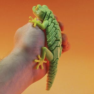 Puede incluir: Una figura de lagarto articulada de color verde claro con un cuerpo texturizado y segmentado y ojos amarillos distintivos. El lagarto presenta un patrón de superficie escamoso detallado en la espalda y las extremidades, con dedos extendidos.
