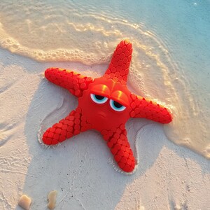 May include: A bright red, five-armed starfish toy with a textured, scale-like surface pattern. The starfish features two large, expressive eyes with white sclera, blue irises, and black pupils, giving it a distinct, animated facial expression.